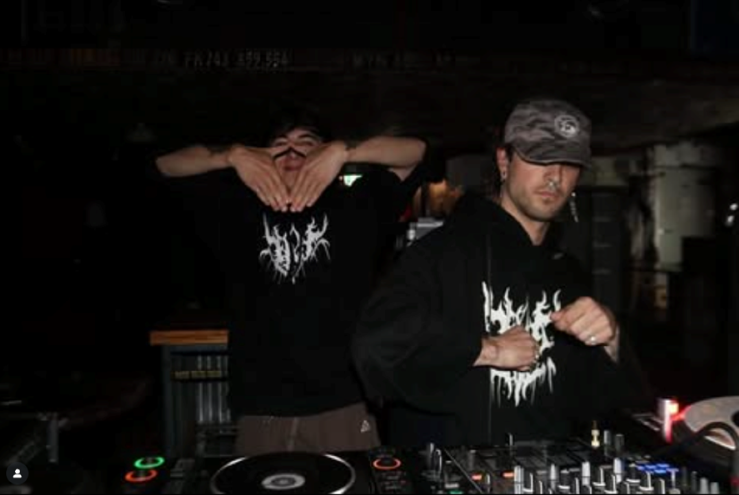 Two people in DNG tees, standing behind the decks - one has their hands in a triangle in front of their face